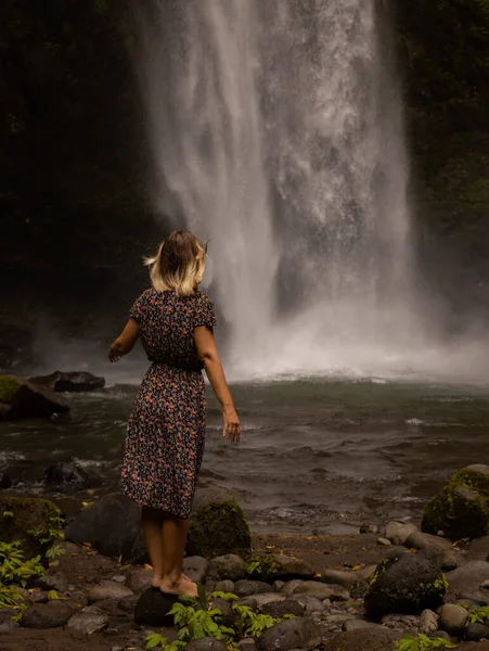 Caucasian woman enjoying waterfall landscape. Nature and environment concept. Travel lifestyle. Woman wearing dress. View from back. Copy space. Nung Nung waterfall in Bali, Indonesia