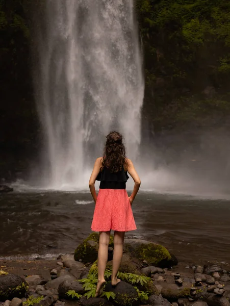 Traveler woman enjoying waterfall landscape. Caucasian woman wearing pink dress. Nature and environment concept. Travel lifestyle. View from back. Copy space. Nung Nung waterfall in Bali, Indonesia