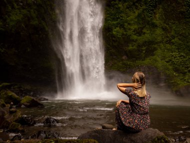 Genç beyaz kadın kayada oturuyor ve şelale manzarasının tadını çıkarıyor. Suyun enerjisi. Seyahat yaşam tarzı. Arkadan bak. Nung Nung Şelalesi, Bali, Endonezya