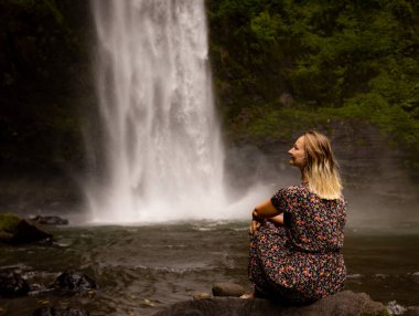 Genç beyaz kadın kayada oturuyor ve şelale manzarasının tadını çıkarıyor. Suyun enerjisi. Seyahat yaşam tarzı. Arkadan bak. Nung Nung Şelalesi, Bali, Endonezya
