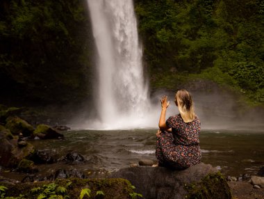 Genç beyaz kadın kayada oturuyor ve şelale manzarasının tadını çıkarıyor. Suyun enerjisi. Seyahat yaşam tarzı. Arkadan bak. Nung Nung Şelalesi, Bali, Endonezya