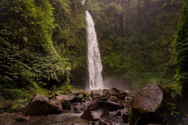 Şelale manzarası. Tropikal yağmur ormanlarında saklı güzel bir şelale. Büyük taşlı ön plan. Yavaş deklanşör hızı, hareket fotoğrafçılığı. Seyahat ve macera. Nung Nung Şelalesi, Bali, Endonezya