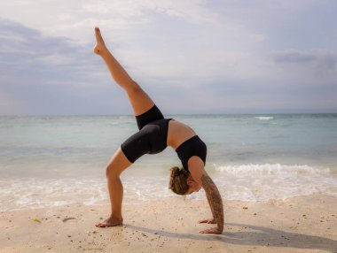 Plaj yogası Genç kadın Eka Pada Chakrasana, Tek Ayaklı Tekerlek Pozu çalışıyor. Yukarı bakan yay pozisyonu derin bir arka virajdır. Esneklik, güç, sabır. Yoga inzivası. Thomas Sahili, Bali, Endonezya