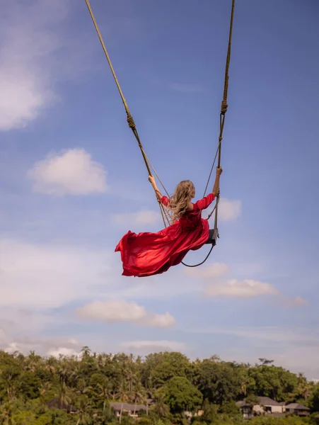 Bali swing trend. Caucasian woman in long red dress swinging in the jungle rainforest. Vacation in Asia. Travel lifestyle. Blue sky with white clouds. View from back. Bongkasa, Bali, Indonesia