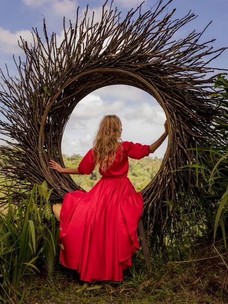 Bali trend. Straw nest in tropical forest. Caucasian woman in long red dress taking photo in a straw nest. Vacation in Asia. Travel lifestyle. Summer concept. View from back. Bongkasa, Bali, Indonesia