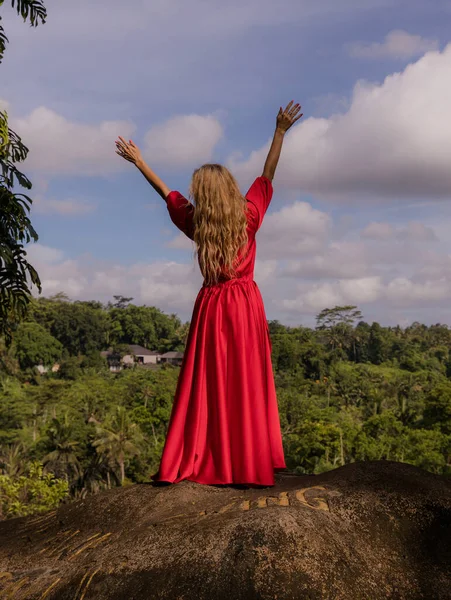Bali trend photo. Caucasian woman in long red dress standing on big stone in tropical rainforest. Vacation in Asia. Travel lifestyle. Summer concept. View from back. Bongkasa, Bali, Indonesia