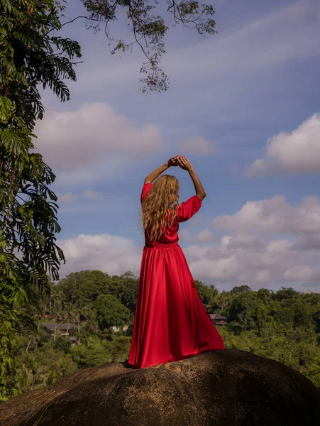 Bali trend photo. Caucasian woman in long red dress standing on big stone in tropical rainforest. Vacation in Asia. Travel lifestyle. Summer concept. View from back. Bongkasa, Bali, Indonesia