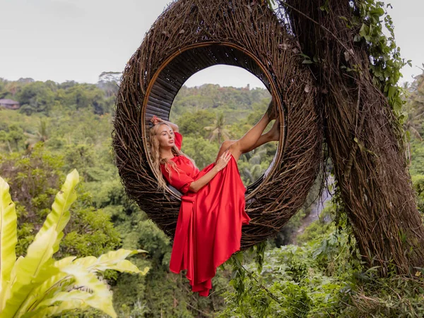 Bali trend. Straw nest in tropical forest. Caucasian woman in long red dress taking photo in a straw nest. Vacation in Asia. Travel lifestyle. Summer concept. Bongkasa, Bali, Indonesia