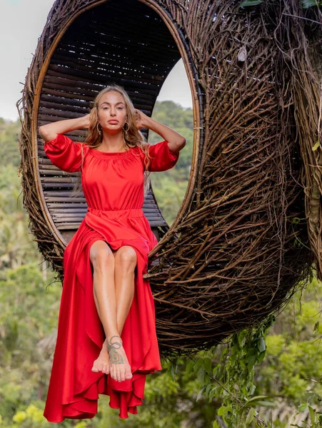 Bali trend. Straw nest in tropical forest. Caucasian woman in long red dress taking photo in a straw nest. Vacation in Asia. Travel lifestyle. Summer concept. Bongkasa, Bali, Indonesia