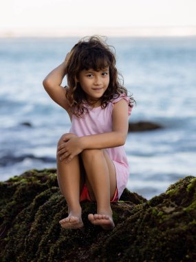Genç kız okyanusun yakınındaki kayada oturuyor. Gülümseyen kız. Mutlu bir çocukluk. Kumsalda vakit geçirmek. Asya 'da tatil. Yaz kavramı. Pandawa Plajı, Bali, Endonezya