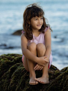 Genç kız okyanusun yakınındaki kayada oturmuş doğanın tadını çıkarıyor. Mutlu bir çocukluk. Kumsalda vakit geçirmek. Asya 'da tatil. Yaz kavramı. Pandawa Plajı, Bali, Endonezya