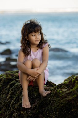 Genç kız okyanusun yakınındaki kayada oturmuş doğanın tadını çıkarıyor. Mutlu bir çocukluk. Kumsalda vakit geçirmek. Asya 'da tatil. Yaz kavramı. Pandawa Plajı, Bali, Endonezya