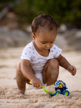Bebek kumlu sahilde oturuyor, kum kepçesi ve oyuncak arabayla oynuyor. Sıcak, güneşli bir gün. Mutlu bir çocukluk. Sevimli küçük çocuk. Denizde yaz tatili. Pandawa Plajı, Bali, Endonezya