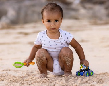 Bebek kumlu sahilde oturuyor, kum kepçesi ve oyuncak arabayla oynuyor. Sıcak, güneşli bir gün. Mutlu bir çocukluk. Sevimli küçük çocuk. Denizde yaz tatili. Pandawa Plajı, Bali, Endonezya