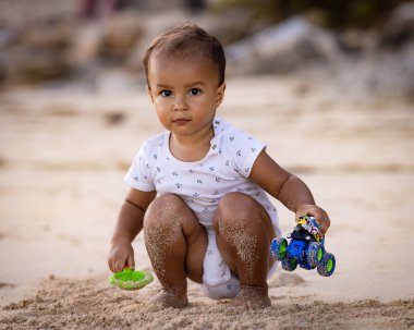 Bebek kumlu sahilde oturuyor, kum kepçesi ve oyuncak arabayla oynuyor. Sıcak, güneşli bir gün. Mutlu bir çocukluk. Sevimli küçük çocuk. Denizde yaz tatili. Pandawa Plajı, Bali, Endonezya