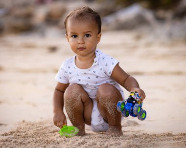 Bebek kumlu sahilde oturuyor, kum kepçesi ve oyuncak arabayla oynuyor. Sıcak, güneşli bir gün. Mutlu bir çocukluk. Sevimli küçük çocuk. Denizde yaz tatili. Pandawa Plajı, Bali, Endonezya