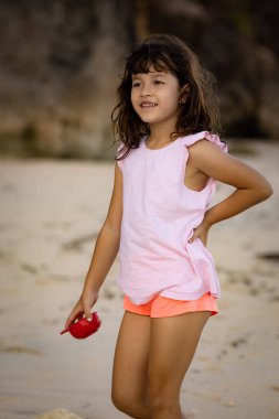 Genç bir kız kumsalda yürüyor, elinde kırmızı bir oyuncak var. Sıcak, güneşli bir gün. Mutlu bir çocukluk. Sevimli küçük kız. Denizde yaz tatili. Okyanus kenarında vakit geçirmek. Pandawa Plajı, Bali, Endonezya