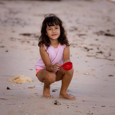 Kumsalda oturan genç bir kız, elinde kırmızı bir oyuncak var. Sıcak, güneşli bir gün. Mutlu bir çocukluk. Sevimli küçük kız. Denizde yaz tatili. Okyanus kenarında vakit geçirmek. Pandawa Plajı, Bali, Endonezya
