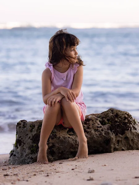 Genç kız okyanusun yakınındaki kayada oturmuş doğanın tadını çıkarıyor. Mutlu bir çocukluk. Kumsalda vakit geçirmek. Asya 'da tatil. Yaz kavramı. Pandawa Plajı, Bali, Endonezya