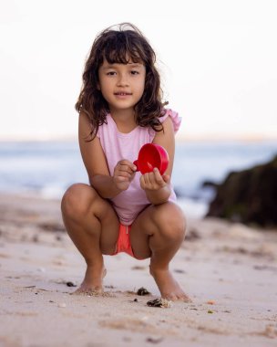 Kumsalda oturan genç bir kız, elinde kırmızı bir oyuncak var. Sıcak, güneşli bir gün. Mutlu bir çocukluk. Sevimli küçük kız. Denizde yaz tatili. Okyanus kenarında vakit geçirmek. Pandawa Plajı, Bali, Endonezya