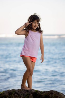 Okyanusun yanındaki kayanın üzerinde duran genç kız. Komik surat ifadesi. Mutlu bir çocukluk. Kumsalda vakit geçirmek. Asya 'da tatil. Yaz kavramı. Pandawa Plajı, Bali, Endonezya