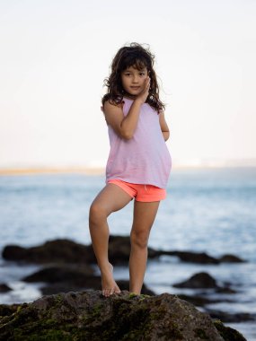 Genç kız okyanusun yanındaki kayanın üzerinde duruyor ve gülümsüyor. Mutlu bir çocukluk. Kumsalda vakit geçirmek. Sevimli küçük kız. Asya 'da tatil. Yaz kavramı. Pandawa Plajı, Bali, Endonezya
