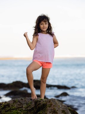 Genç kız okyanusun yanındaki kayanın üzerinde duruyor ve gülümsüyor. Mutlu bir çocukluk. Kumsalda vakit geçirmek. Sevimli küçük kız. Asya 'da tatil. Yaz kavramı. Pandawa Plajı, Bali, Endonezya