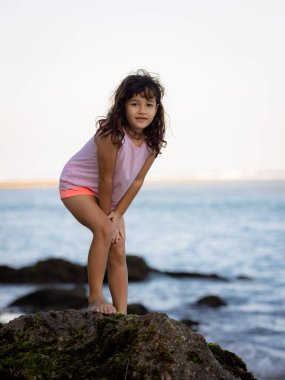 Genç kız okyanusun yanındaki kayanın üzerinde duruyor ve gülümsüyor. Mutlu bir çocukluk. Kumsalda vakit geçirmek. Sevimli küçük kız. Asya 'da tatil. Yaz kavramı. Pandawa Plajı, Bali, Endonezya