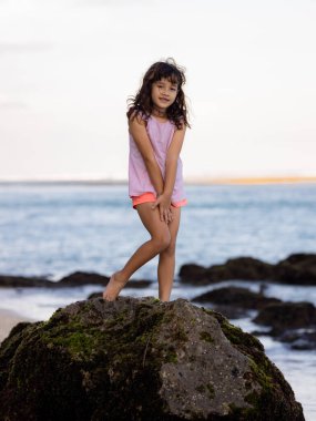 Genç kız okyanusun yanındaki kayanın üzerinde duruyor ve gülümsüyor. Mutlu bir çocukluk. Kumsalda vakit geçirmek. Sevimli küçük kız. Asya 'da tatil. Yaz kavramı. Pandawa Plajı, Bali, Endonezya