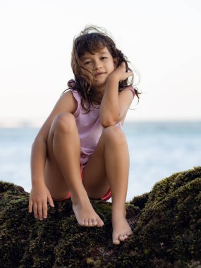 Genç kız okyanusun yakınındaki kayada oturuyor ve gülümsüyor. Mutlu bir çocukluk. Kumsalda vakit geçirmek. Sevimli küçük kız. Asya 'da tatil. Yaz kavramı. Pandawa Plajı, Bali, Endonezya