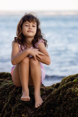 Genç kız okyanusun yakınındaki kayada oturuyor ve gülümsüyor. Mutlu bir çocukluk. Kumsalda vakit geçirmek. Sevimli küçük kız. Asya 'da tatil. Yaz kavramı. Pandawa Plajı, Bali, Endonezya