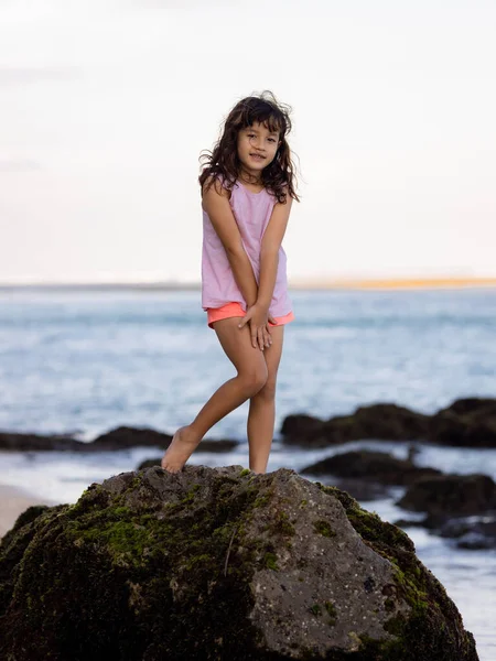 Genç kız okyanusun yanındaki kayanın üzerinde duruyor ve gülümsüyor. Mutlu bir çocukluk. Kumsalda vakit geçirmek. Sevimli küçük kız. Asya 'da tatil. Yaz kavramı. Pandawa Plajı, Bali, Endonezya
