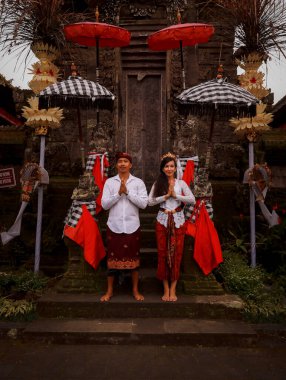 Bali 'deki tapınağın yanında duran çok kültürlü bir çift. Eller namaste 'de. Beyaz kadın ve Balili koca geleneksel kıyafetler giyiyor. Penjor bambu ve şemsiye süslemesi. Penglipuran, Bali