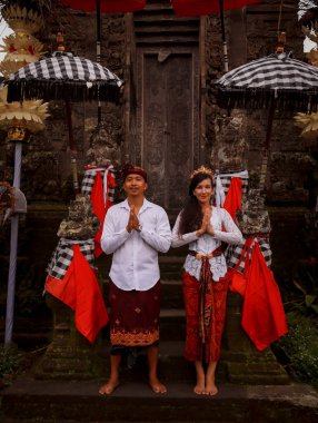 Bali 'deki tapınağın yanında duran çok kültürlü bir çift. Eller namaste 'de. Beyaz kadın ve Balili koca geleneksel kıyafetler giyiyor. Penjor bambu ve şemsiye süslemesi. Penglipuran, Bali