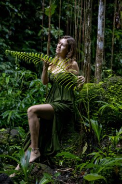 Ormandaki bir kadın yemyeşil bitkilerle çevrili bir taşın üzerinde oturmuş eğreltiotu yaprağına dokunuyor. Tropikal yağmur ormanlarında yeşil elbiseli beyaz bir kadın. Doğa konsepti. Asya 'ya git. Bali, Endonezya