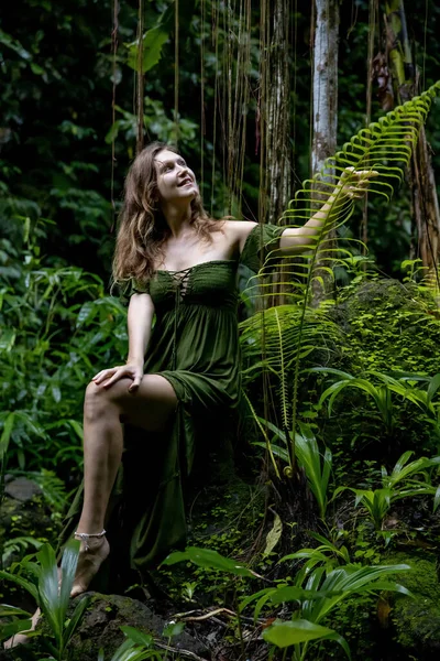 ジャングルの中の女性の石緑豊かな緑の植物に囲まれ シダの葉に触れて座っている 熱帯雨林の白人女性は 緑のドレスを着ている 自然概念 アジアへの旅行 インドネシアのバリ — ストック写真