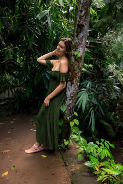 Beyaz kadın tropikal yağmur ormanlarında ağaca yaslanıyor. Uzun yeşil elbise giyen güzel bir kadın. Ormanda yürüyüş yolu. Doğa konsepti. Asya 'ya git. Bali Adası, Endonezya