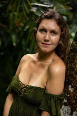 Tropikal yağmur ormanlarında bir ağaca yaslanan beyaz kadın portresi. Yeşil elbise giyen güzel bir kadın. Doğa konsepti. Asya 'ya git. Bali Adası, Endonezya