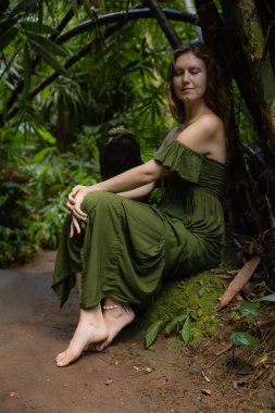 Ormandaki kadın bambunun altında bir taşın üzerinde oturuyor. Kapalı gözler. Tropikal yağmur ormanlarında yeşil elbiseli beyaz bir kadın. Doğa konsepti. Asya 'ya git. Bali Adası, Endonezya