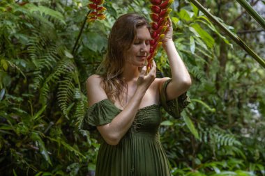 İki kırmızı sarmal çiçekli güzel bir kadının portresi. Kapalı gözler. Tropikal yağmur ormanlarında yeşil elbiseli beyaz bir kadın. Doğa ve çevre kavramı. Asya 'ya git. Bali, Endonezya
