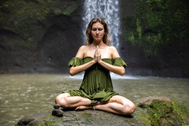 Yoga nilüfer pozu. Genç beyaz kadın taşın üzerinde oturuyor, meditasyon yapıyor, yoga yapıyor, şelalede pranayama yapıyor. Eller namaste mudra 'da. Kapalı gözler. Yoga inzivası. Tibumana Şelalesi, Bali, Endonezya