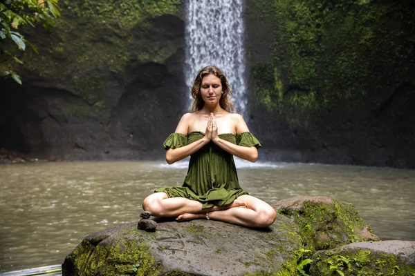 Yoga nilüfer pozu. Genç beyaz kadın taşın üzerinde oturuyor, meditasyon yapıyor, yoga yapıyor, şelalede pranayama yapıyor. Eller namaste mudra 'da. Kapalı gözler. Yoga inzivası. Tibumana Şelalesi, Bali, Endonezya