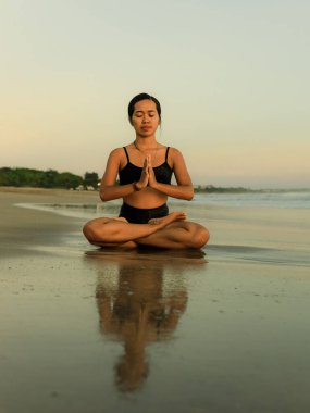 Sahilde yoga antrenmanı. Lotus pozu. Padmasana. Eller namaste mudra 'da. Kapalı gözler. Meditasyon ve konsantrasyon. Zen hayatı. Bedensel ve zihinsel rahatlama. Yoga inzivası. Gün batımı zamanı. Seminyak, Bali