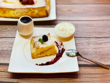 Beyaz tabakta kremalı güveç. Kahvaltıda süzme peynirli tatlı bir dilim turta. Masada tatlı olarak beyaz tabak ve bir çay kaşığı.
