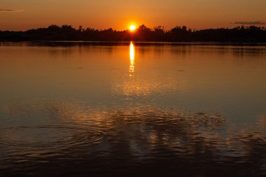 Sakin bir gölde güneşli, renkli bir gün batımı. Güneş suyun yüzeyine yansıyor..