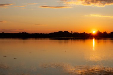 Sakin bir gölde güneşli, renkli bir gün batımı. Güneş suyun yüzeyine yansıyor..