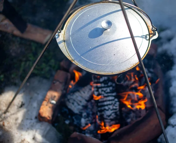 Over the fire hangs a pot in which to cook food. On a hook on a tripod, steam comes out of the pan. Winter Camping outdoor cooking
