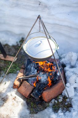 Ateşin üzerinde yemek pişirmek için bir tencere asılı. Üç ayaklı bir kancada, tavadan buhar çıkıyor. Kış kampı açık hava yemekleri.