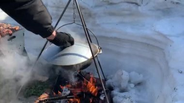 Bir turist tenceresi kışın kamp ateşinin başında durur ve kışın doğada ateş yakar. Adam kazan kapağını açıyor, 4K