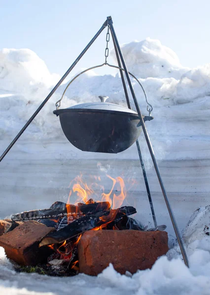 Over the fire hangs a pot in which to cook food. On a hook on a tripod, steam comes out of the pan. Winter Camping outdoor cooking